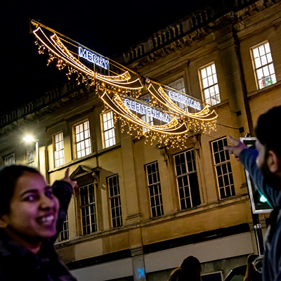 Couple point towards illuminated holiday decorations hanging above a street at night that read 