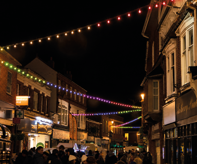 A street scene at night features illuminated decorative illuminated arches in white and warm white near the entrance of Exeter Central station.