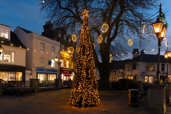 Illuminated cone tree with a star in centre of town square surrounded by festoon strung between lantern style lampposts.
