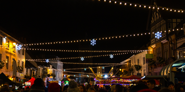 Warm white festoon lights are hanging across a narrow outdoor shopping street between buildings.