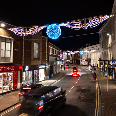 A nighttime street scene with cars driving under festive blue and white holiday motifs lights strung across the road between buildings.