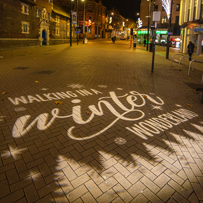A quiet city street at night is decorated with illuminated holiday lights hanging above and along the buildings, with a 'walking in a winter wonderland' design projected onto the paved walkway.