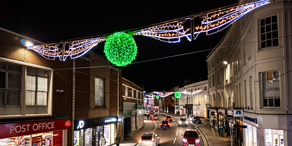 A nighttime street scene with festive holiday cross street lights featuring green illuminated spheres and kinetic motifs hanging above a road lined with shops and cars.