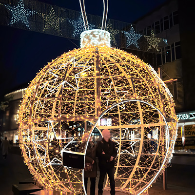 A large illuminated bauble decoration displayed on a street at night with a couple walking through it with star shaped lights in the background.