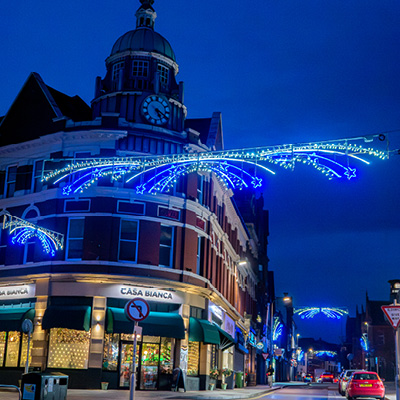 Blue and white illuminated star cross street motifs arching over a crossroads with a historic clock tower set against a deep blue evening sky.