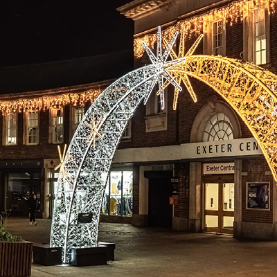 A street scene at night features illuminated decorative illuminated arches in white and warm white near the entrance of Exeter Central station.