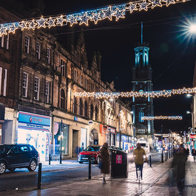 A nighttime street scene with people walking along a sidewalk decorated with hanging star-shaped lights, parked cars, and illuminated shopfronts.