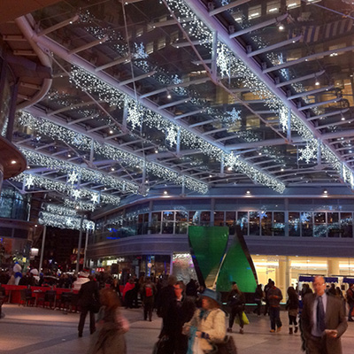 A crowd of people is gathered under a glass ceiling decorated with white icicle lights and hanging illuminated snowflakes in an urban retail environment at night.