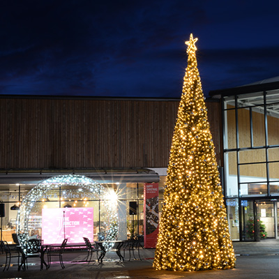 A tall Christmas tree decorated with warm white lights and a star on top stands illuminated in a retail park plaza at night.