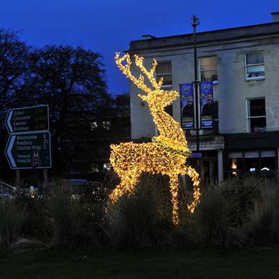 A large illuminated reindeer decoration made of warm white lights stands in the middle of a roundabout at dusk.