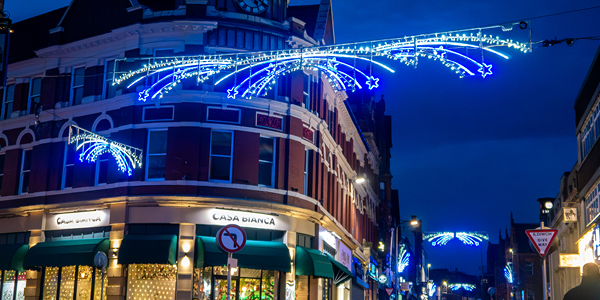 A nighttime street scene shows a building with a clock tower decorated with blue and white star-shaped holiday lights hanging across the street.

