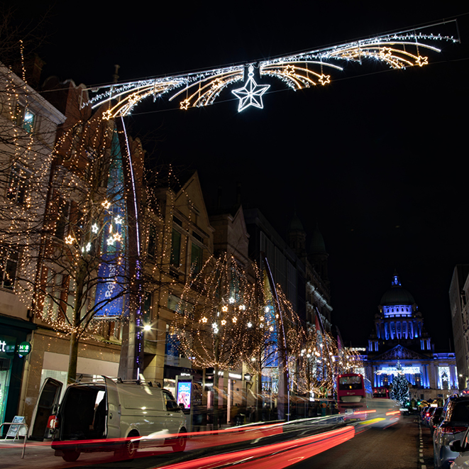Tree lined avenue with star cross street motif, warm white light wrapped trees, with neon stars amongst the trees