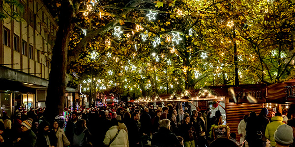 A crowded outdoor Christmas market at night decorated with illuminated star-shaped lights hanging from trees above people browsing wooden stalls.