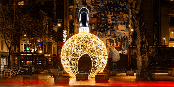 A large Christmas walk-through bauble illuminated in a nighttime urban setting with blurred light trails from passing vehicles and a mural of people on a building wall in the background.