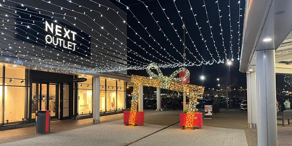 A nighttime outdoor shopping area decorated with white light curtain overhead and a large illuminated gift box archway near the entrance of a store labeled NEXT OUTLET.