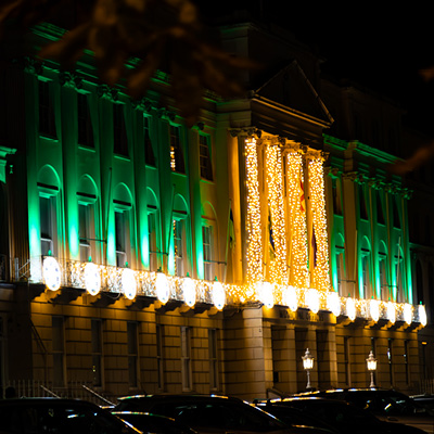 A historic building illuminated at night with green uplighters and warm white lights wrapped around the columns highlighting its architectural details above a row of parked cars.