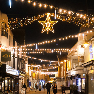 A pedestrian shopping street with overhead warm white festoon interspersed with snowfall tubes and a large hanging star decoration.