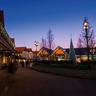 A festive outdoor shopping area at dusk is decorated with string lights, illuminated star-shaped street lamps, and a tall Christmas tree covered in lights.