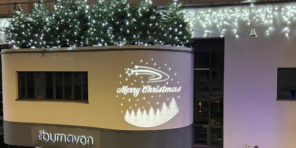 A rooftop decorated with multiple Christmas trees adorned with white string lights and icicle lights hanging along the edge of the building with a bespoke 'Merry Christmas' GOBO projection on the building side.