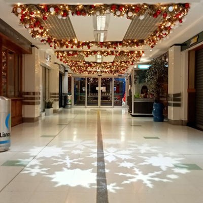 An indoor shopping centre hallway decorated with gold garland with red and gold baubles attached to the ceiling with a snowflake light projection on the floor.