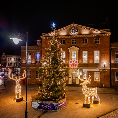 A large Christmas tree decorated with lights and a star on top stands next to a glowing reindeer figure in front of a brick building illuminated at night.