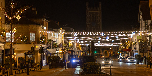 A nighttime street scene decorated with warm white festoon lights and illuminated snowflake ornaments, with cars driving along the road and buildings adorned with festive greenery.