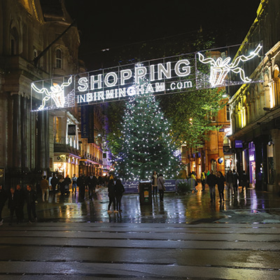 A large Christmas tree decorated with white lights stands in a busy city street beneath a festive illuminated sign that reads 