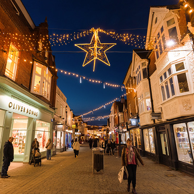 A busy street scene at night shows people walking and sitting in a pedestrian area decorated with festoon, snowfall tubes and illuminated star-shaped ornaments zigzagging along street.