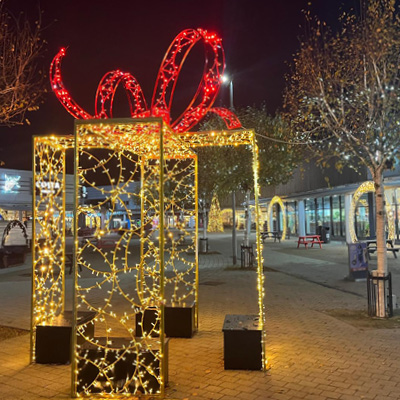 A large outdoor illuminated decorative gift box in warm white with a red light bow on top, set in a nighttime retail park plaza with trees and benches.