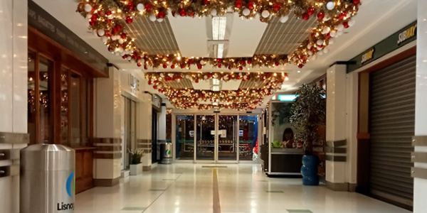 A brightly decorated indoor shopping centre hallway with festive red and gold garland decorated with ornaments hanging from the ceiling.