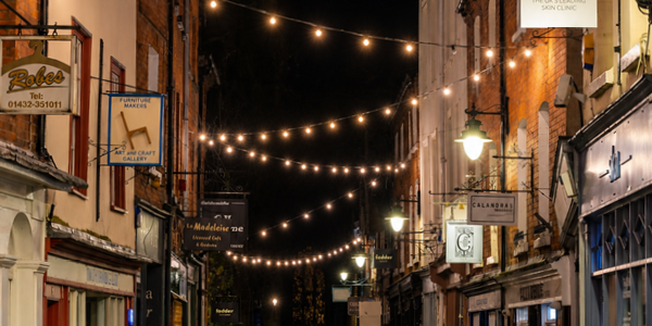A group of people gathered closely together for a festive light switch on, with warm white festoon strung between buildings on a wide street at night.