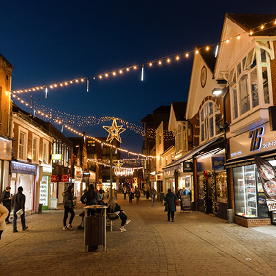 A busy street scene at night shows people walking and sitting in a pedestrian area decorated with festoon, snowfall tubes and illuminated star-shaped ornaments zigzagging along street.