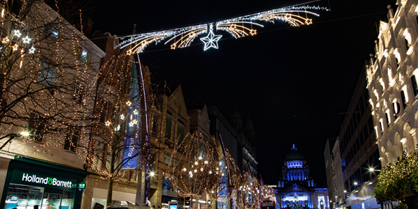 A nighttime street scene decorated with illuminated holiday lights shaped like stars and shooting stars hanging above and wrapped around trees.