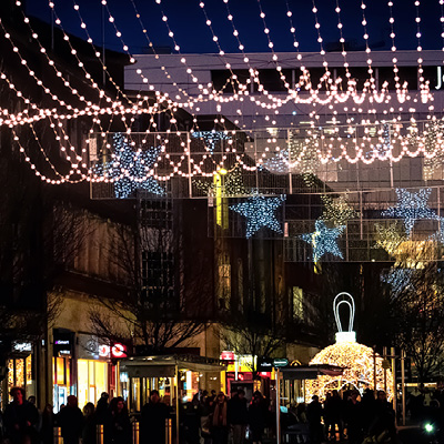 A nighttime street scene with people walking under festive string lights and illuminated star cross street motifs with a giant walk-through bauble in the background.