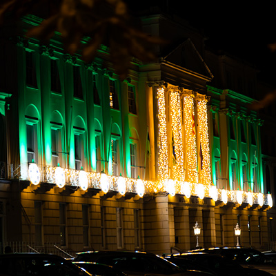 A large building is illuminated at night with green uplighters and yellow motifs, featuring columns wrapped in golden string lights with several parked cars in front.