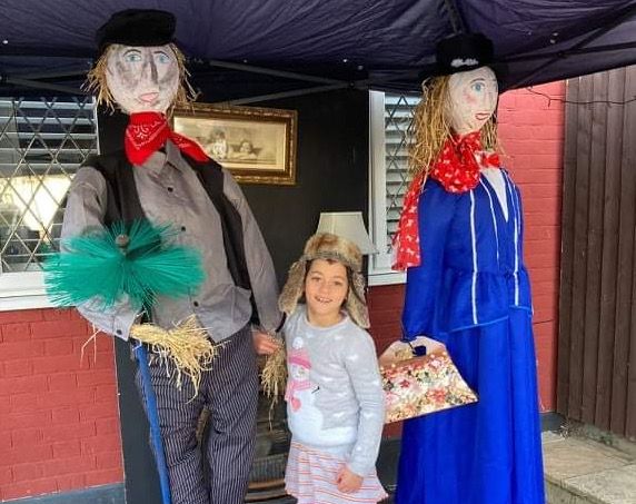 A small girl standing between two scarecrows in front of her house.