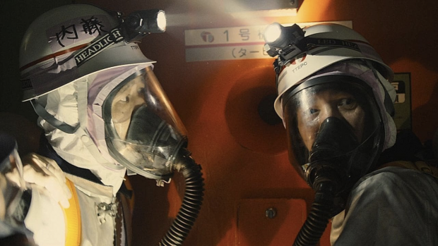 Two workers of the Fukushima 50 are wearing heavy duty protective equipment and oxygen masks. They are in fornt of a nuclear reactor vent. One is looking to the other worker, who is looking away.