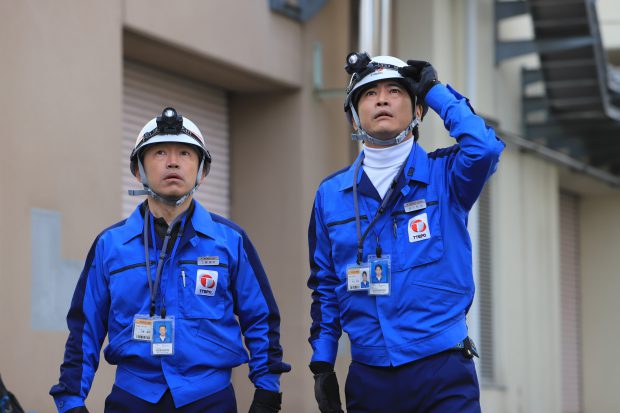Two workers dressed in bright blue uniforms and white helmets gaze up with mouths open and eyes wide open.