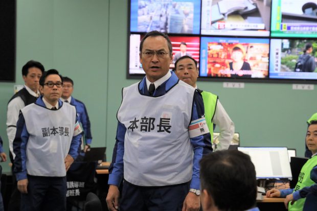 Actor Ken Watanabe stands in the centre of a busy room, mid sentence, mouth open and eyebrows furrowed. There are multiple tv news screens behind him.