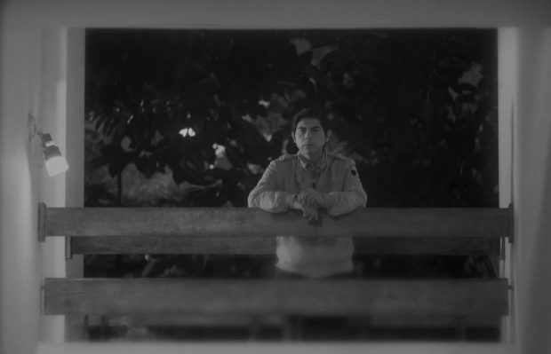Black and white still from Song Without A Name. A man (Párraga) leans on a railing. He has a cigarette in his left hand.
