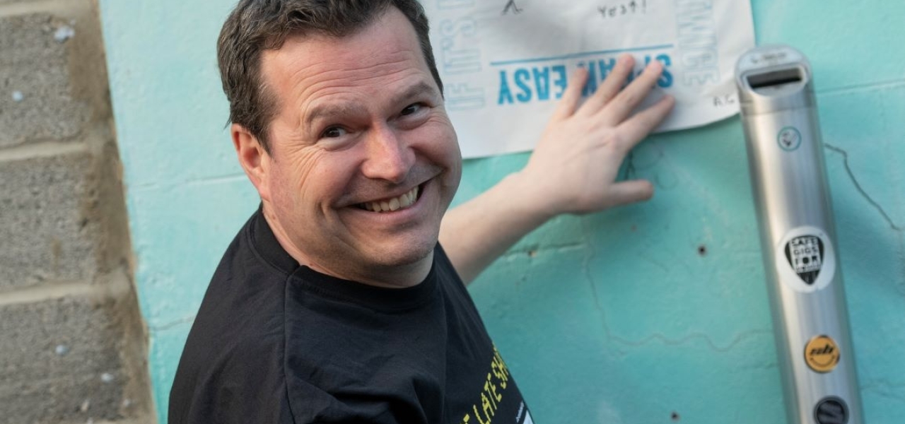 Photo of a male pasting a protest poster to a wall, looking back to smile at the camera