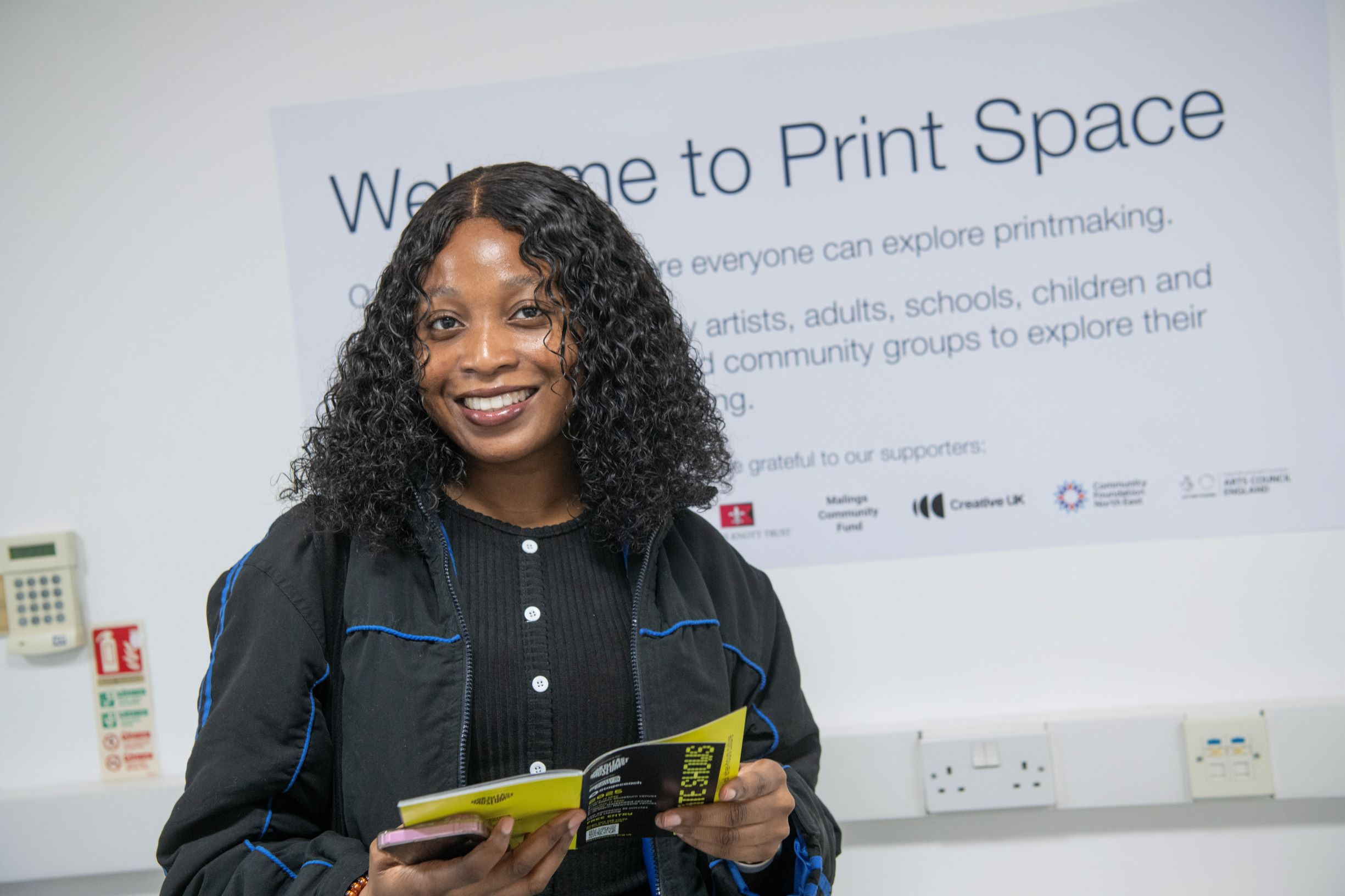 Girl smiling while looking at the brochure - in front of a white wall mural reads 'The Print Space'