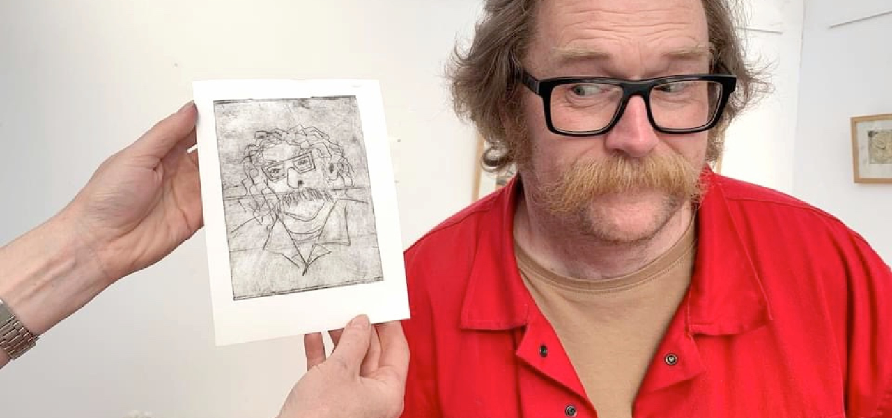 Man with red top, glasses and moustache stands next to a pencil drawing of himself