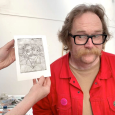 Man with red top, glasses and moustache stands next to a pencil drawing of himself