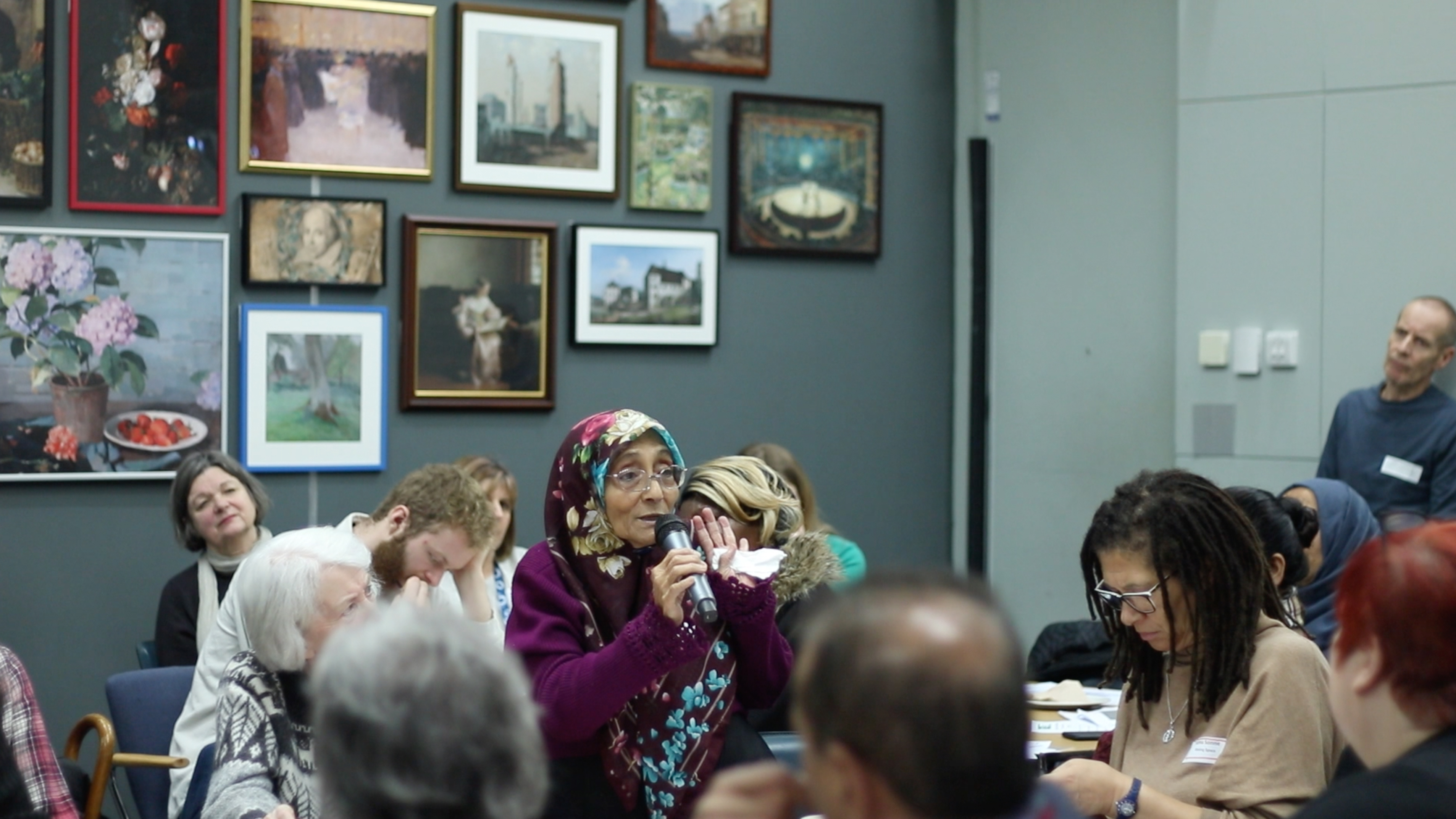 older Asian woman speaking with microphone at event paintings on wall behind