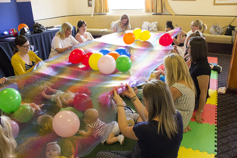 babies sitting on adults laps in a circle arount a big stretched out sheet fiilled with balloons