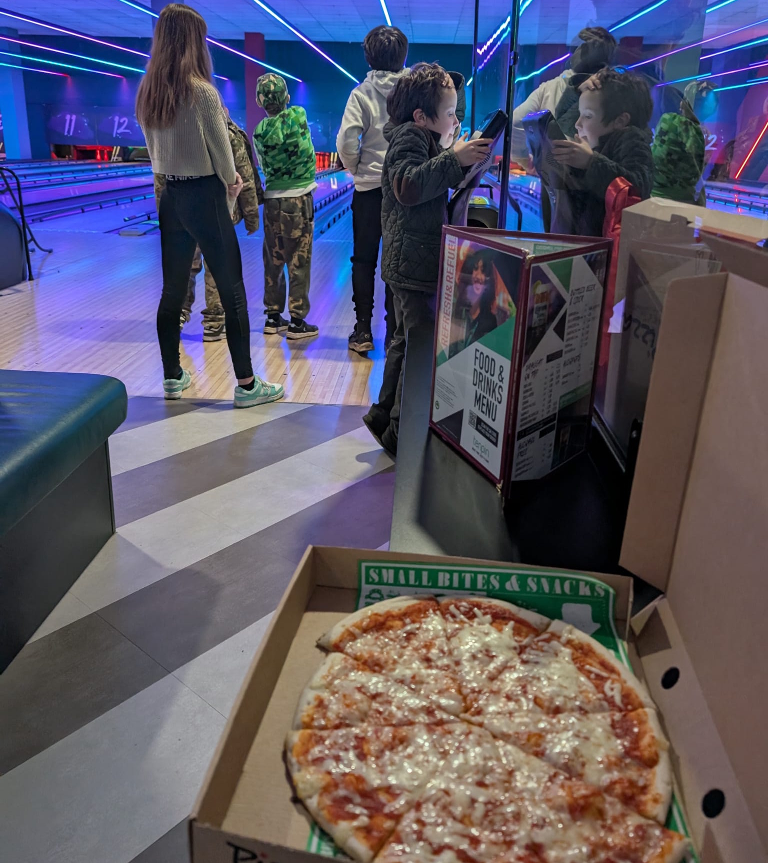 Pizza in a box with a number of children playing pool in the background