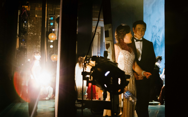 A man and woman dressed in glamorous 1920's clothing grin at each other whilst backstage in a theatre