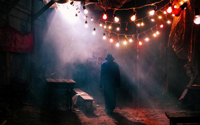 A shadowy figure wearing a hat walks through a darkly lit street with hanging lights above