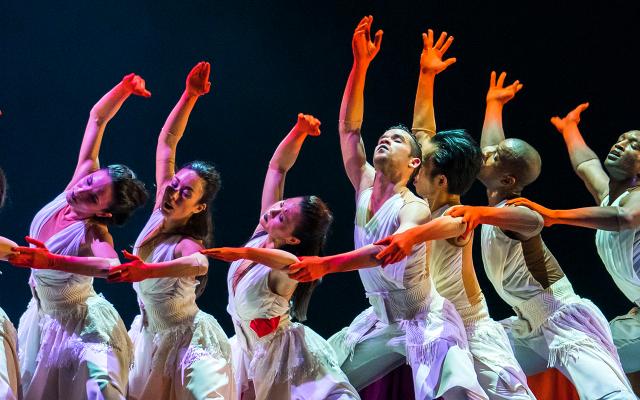 Male and female dancers in white robes with long red gloves each raise a hand aloft against a dark background
